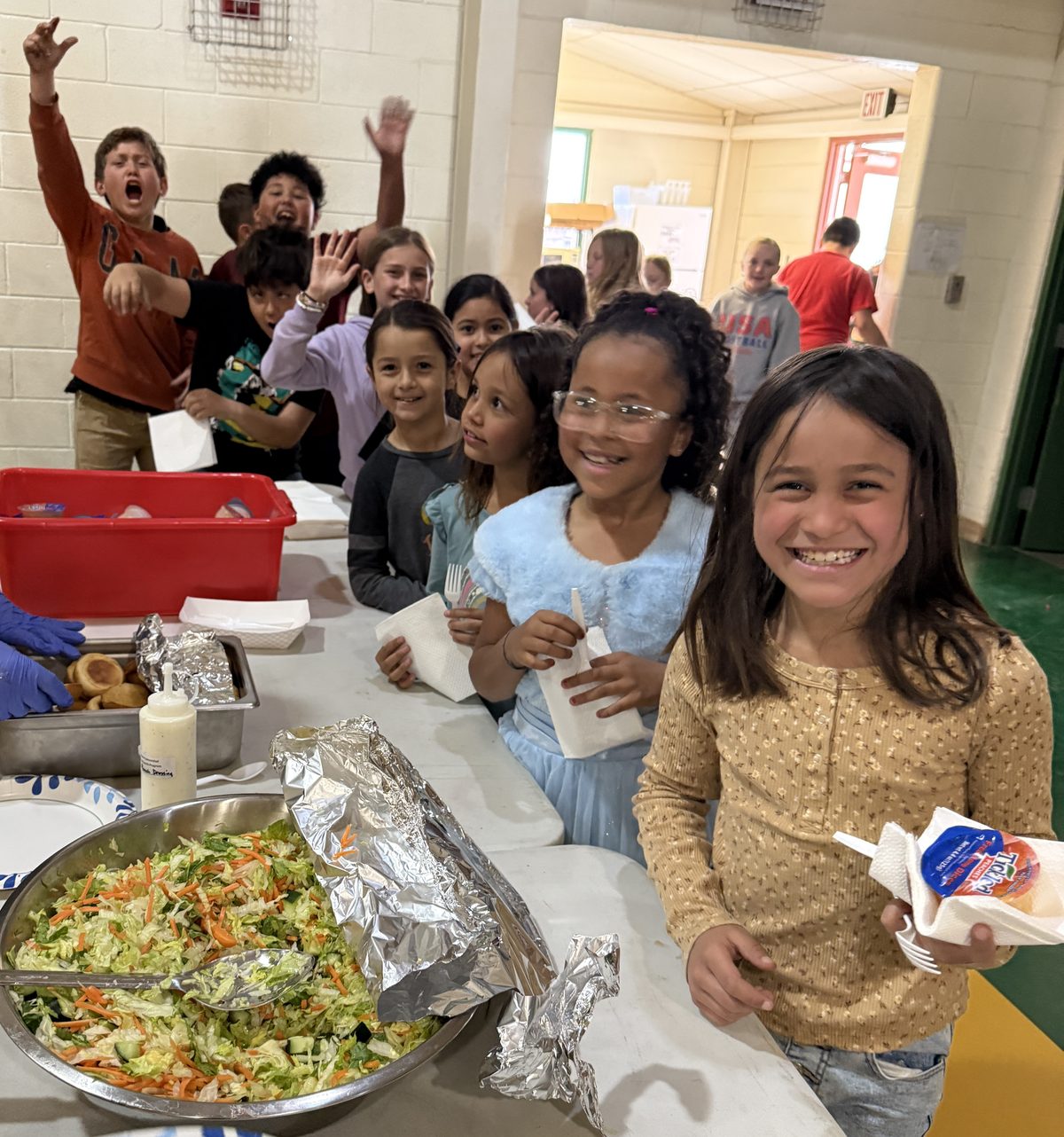 Smiling elementary students in the lunch line with a large bowl of fresh salad in the foreground